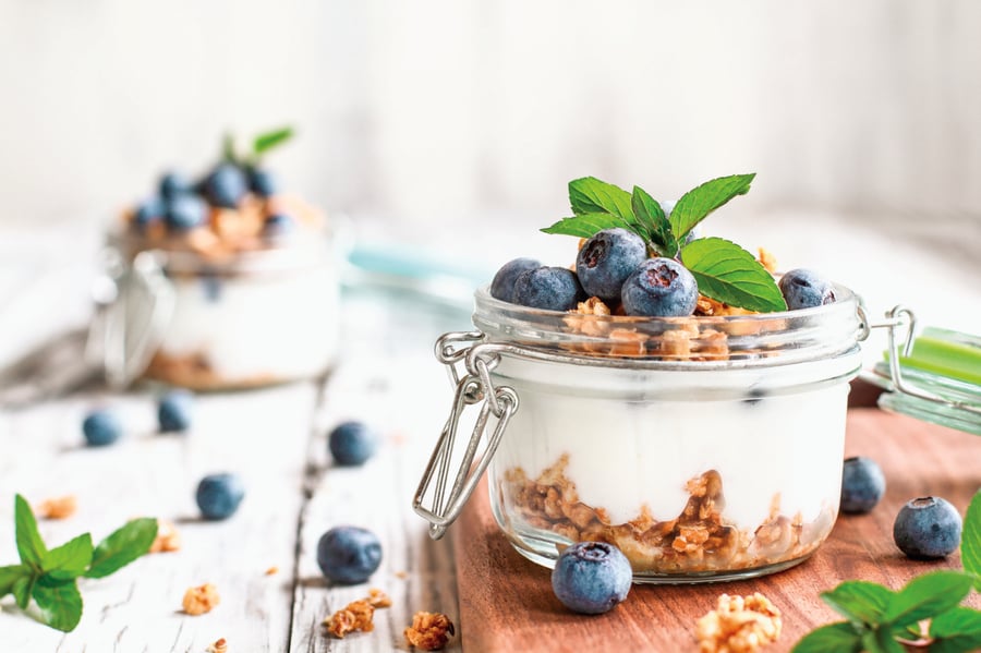 Short jar of vanilla yogurt with granola crumbles and blueberries. Additional granola pieces and blueberries are scattered in the background