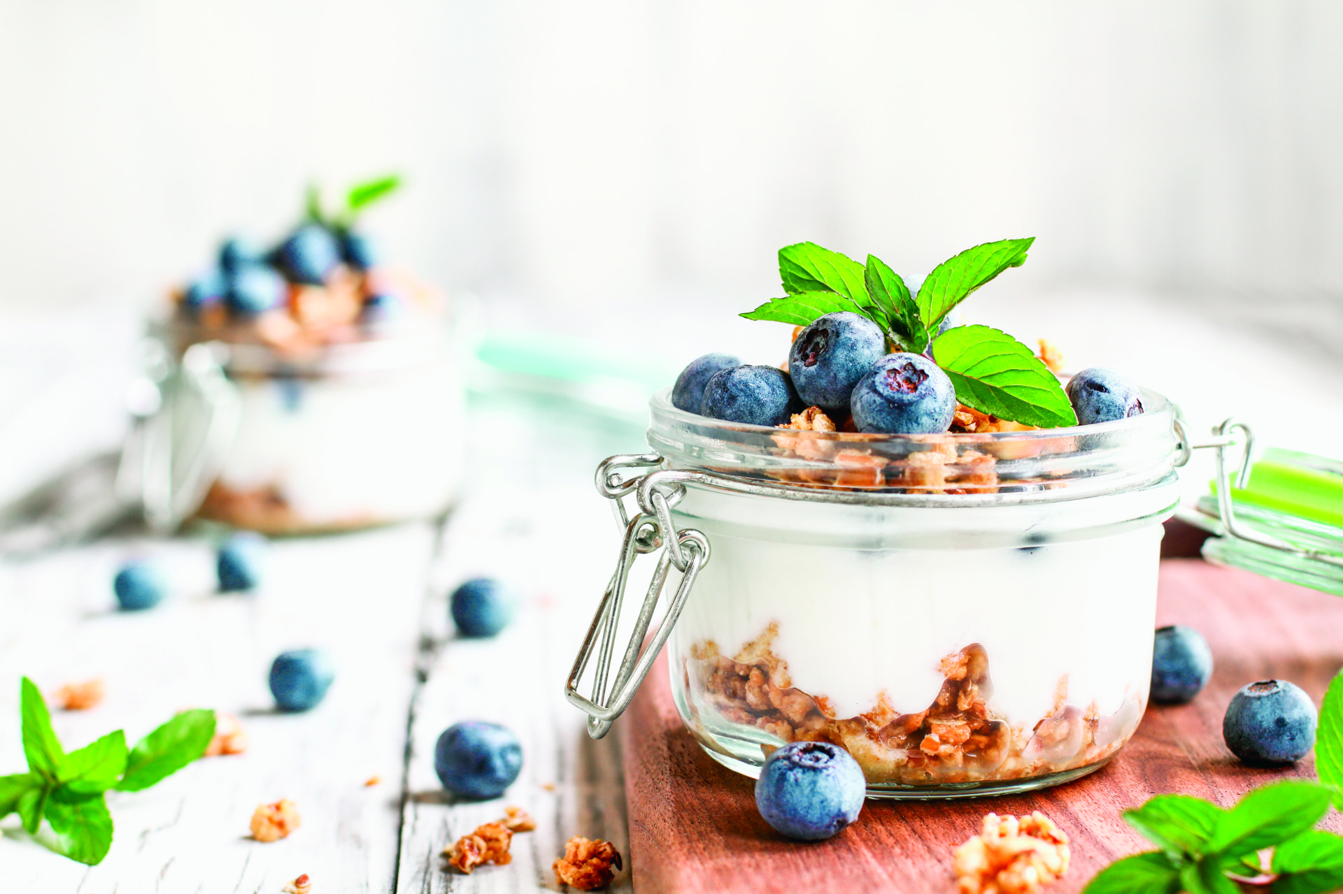 Short jar of vanilla yogurt with granola crumbles and blueberries. Additional granola pieces and blueberries are scattered in the background