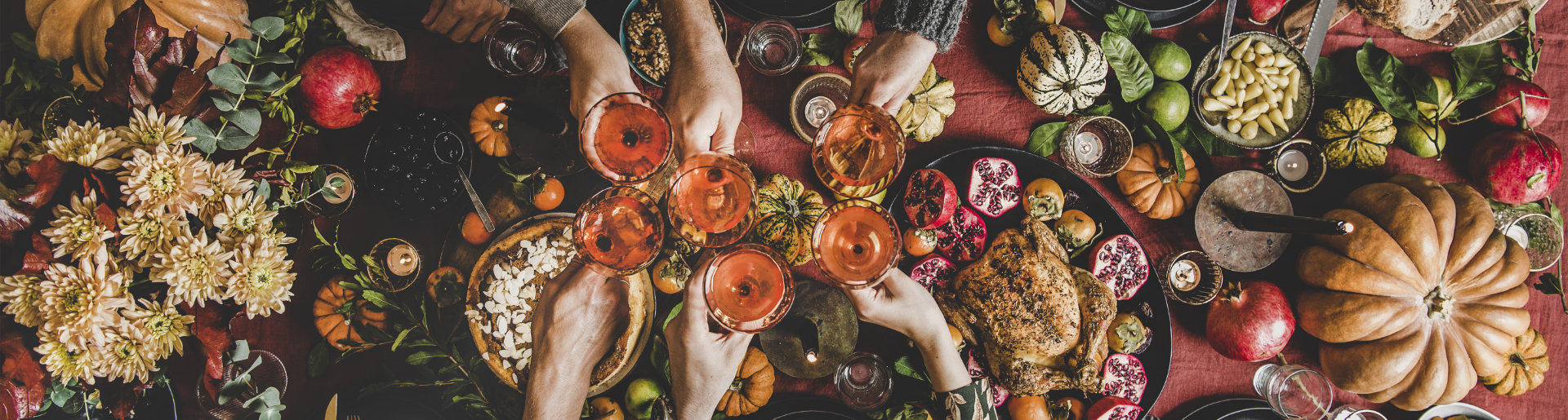 Top down shot of a festive fall gathering, featuring people bringing their cocktail glasses together to cheers. Around their hands you can see pumpkins and pomegranates 